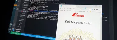 A picture of a computer screen with VSCode opened on a ruby file. On top of this window, there's a smaller window with 'localhost:3000' as url. The website says 'Yay! You're on Rails!' with the ruby on rails logo above it.