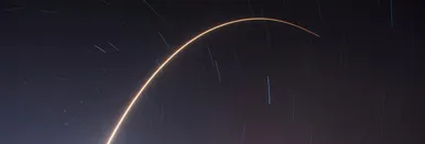 Long exposure photo of a rocket shooting into the night sky, forming a glowing arc over the horizon